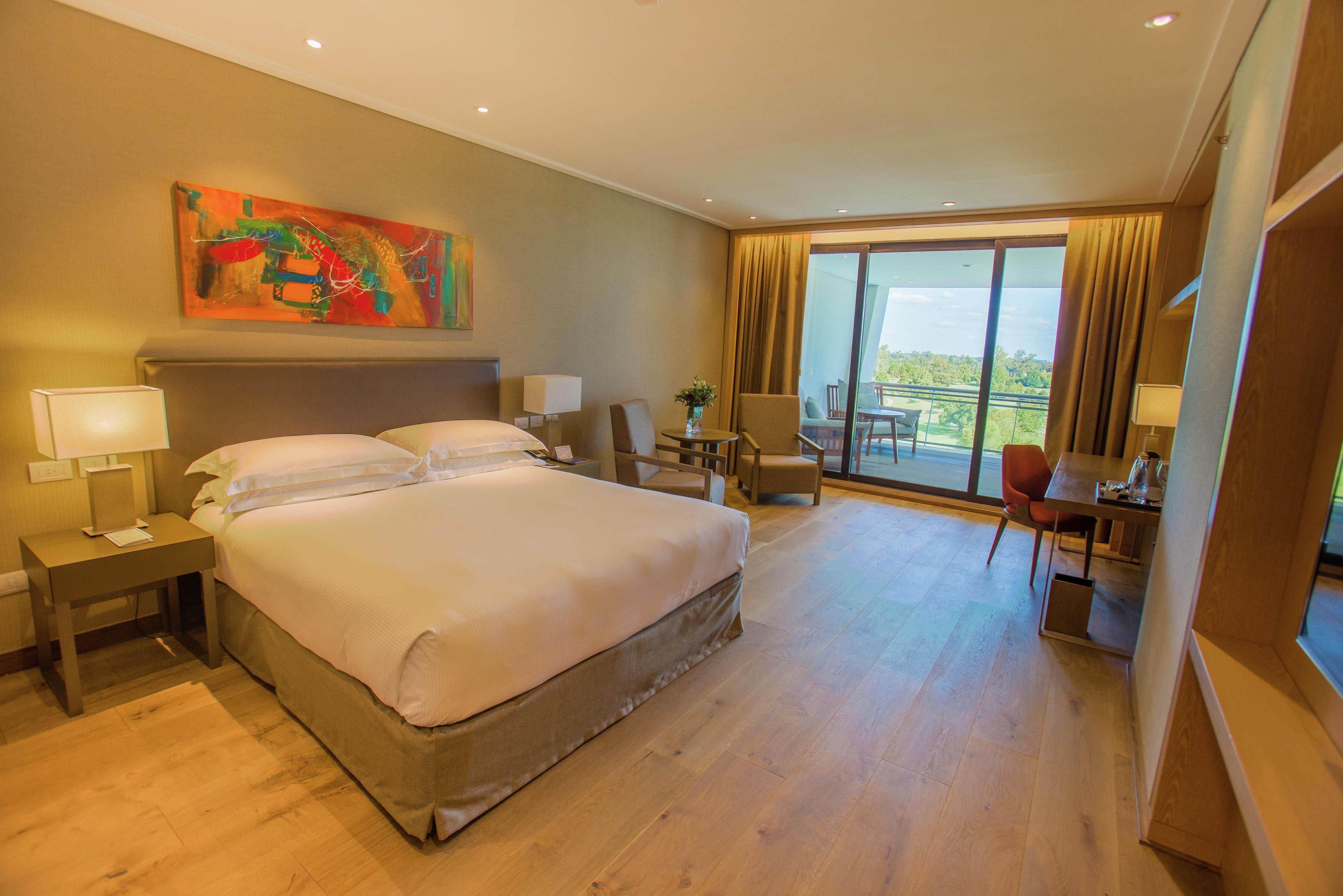 A hotel room with a large bed, a desk with a chair, a small table with a chair, a painting on the wall, and a balcony with a view.