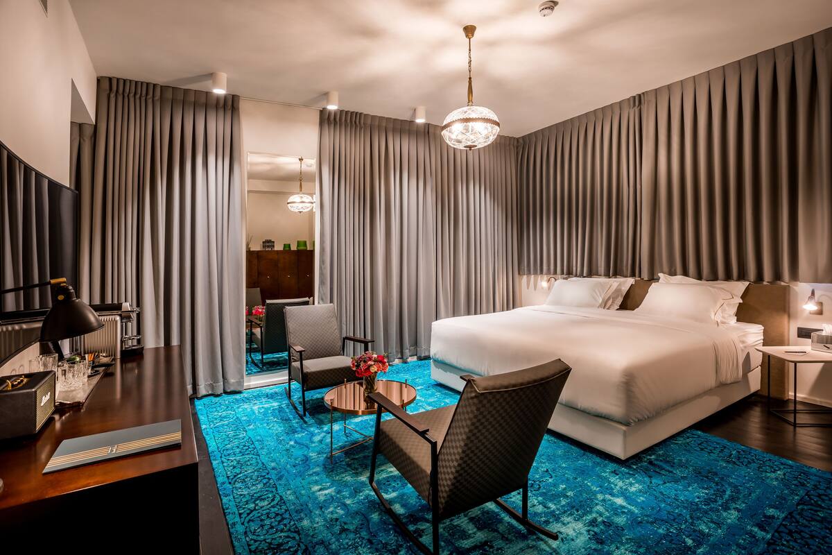 A hotel room with a large bed, a desk with a lamp, a chair, a small table with a vase, and a blue patterned rug.