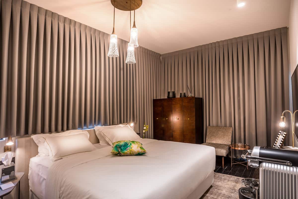 A modern hotel room with a large bed, a wooden wardrobe, a seating area, and a vanity with a mirror.