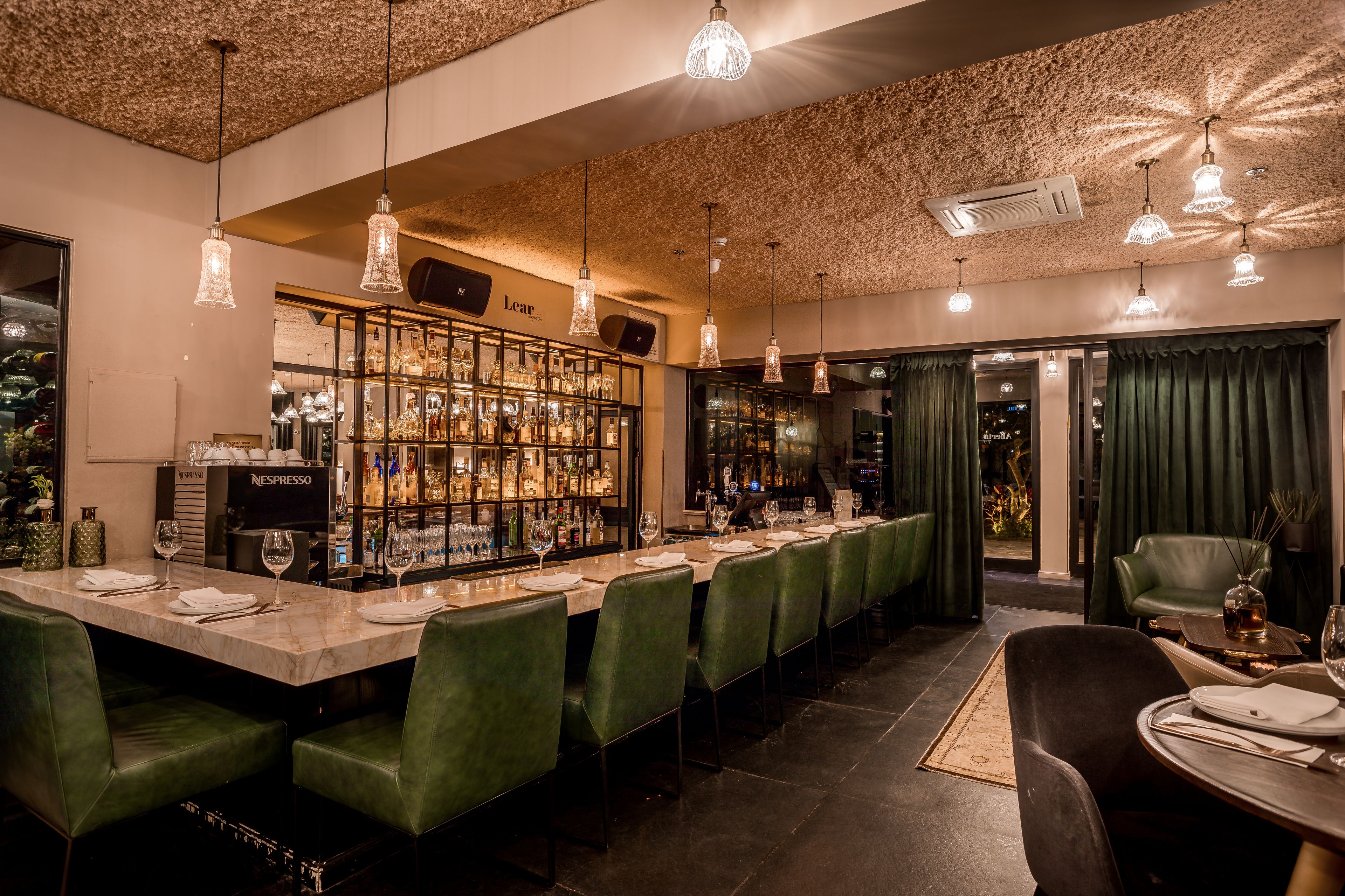 A bar area with a long counter, green upholstered chairs, and pendant lighting.