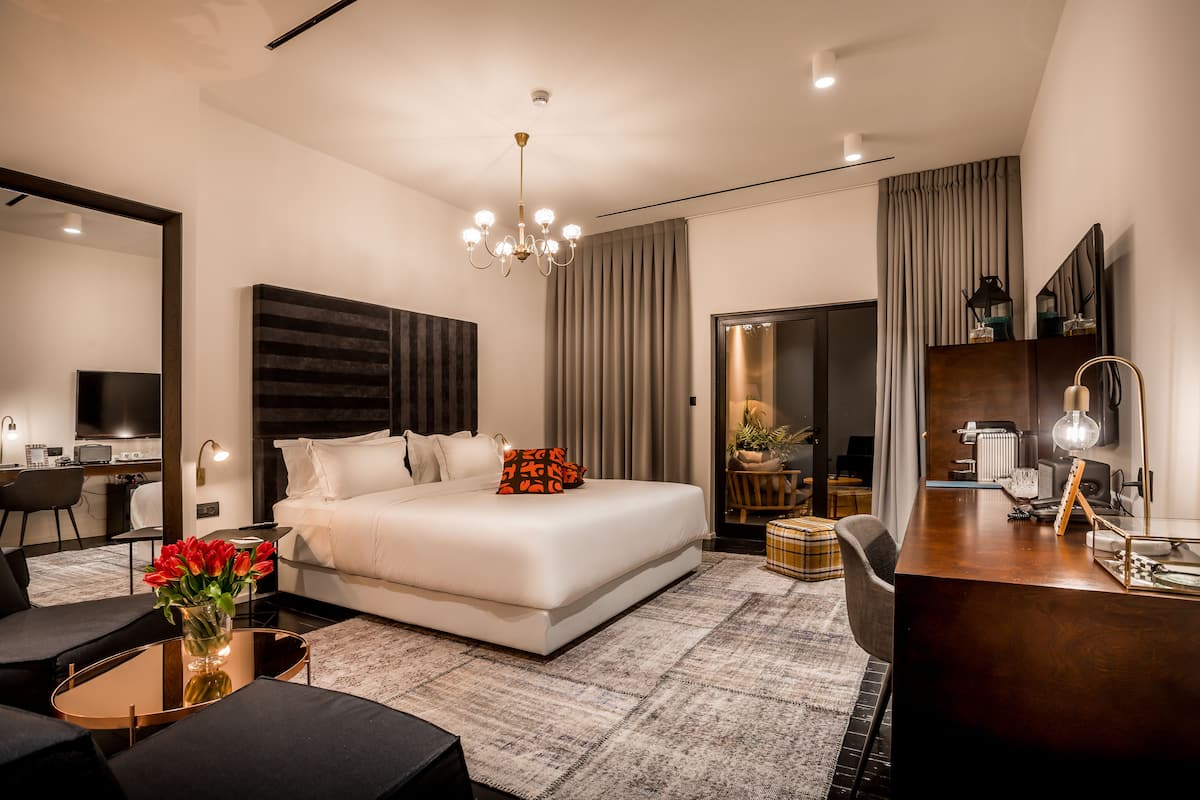 A modern hotel room with a large bed, a desk, a chair, a coffee table, and a vase of flowers.