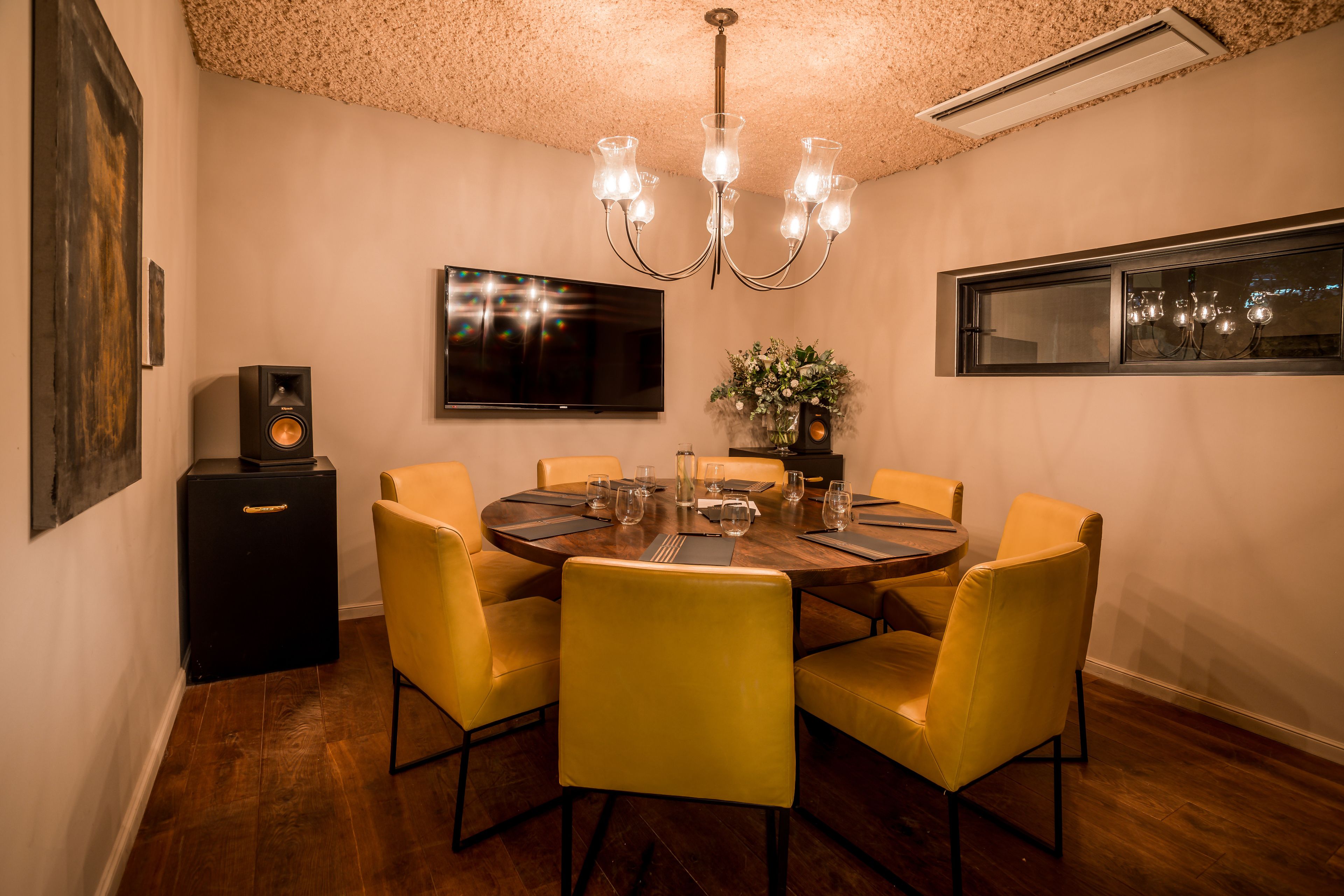 A dining area with a round table, yellow chairs, a chandelier, a flat-screen TV, and a small speaker.