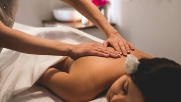 A person receiving a massage on a bed in a room with a white wall and a small table with a vase of flowers in the background.