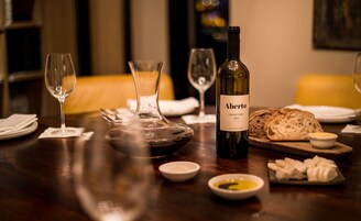 Una mesa de comedor con una botella de vino, copas, pan y recipientes pequeños de condimentos.
