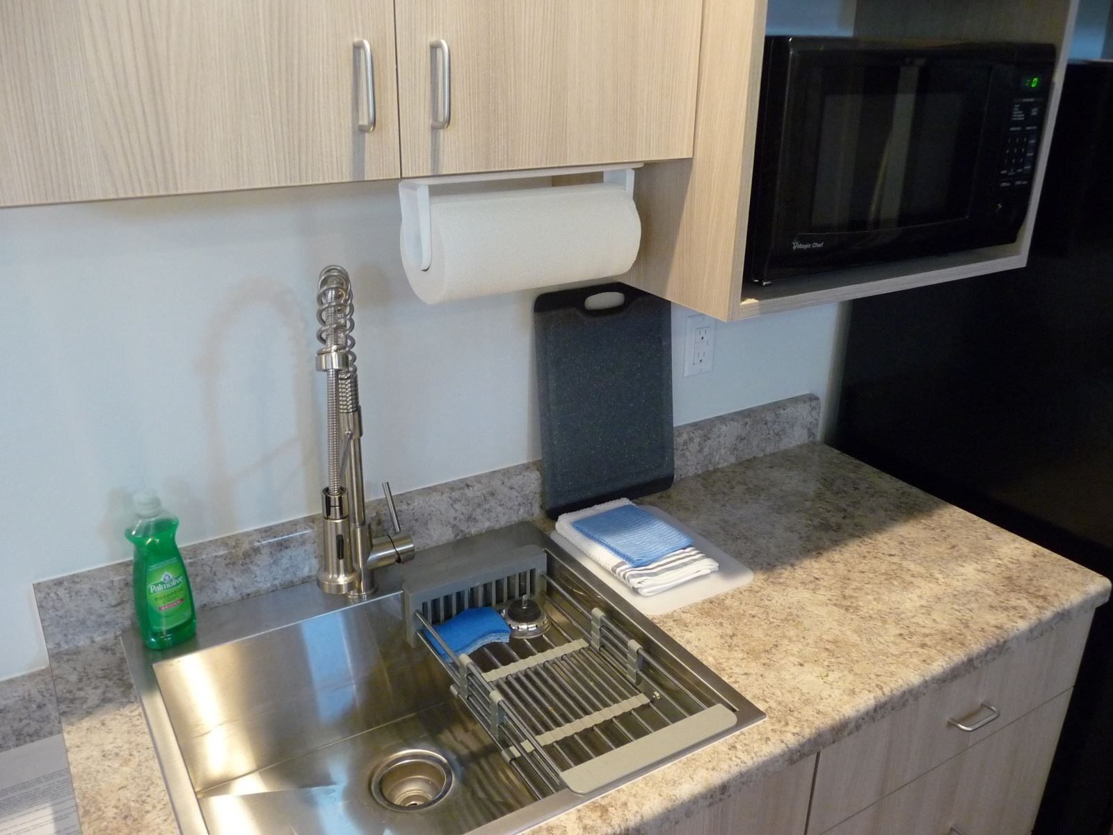 Una cocina con un lavamanos de acero inoxidable, un escurreplatos, una botella de detergente, un dispensador de toallas de papel y un microondas.