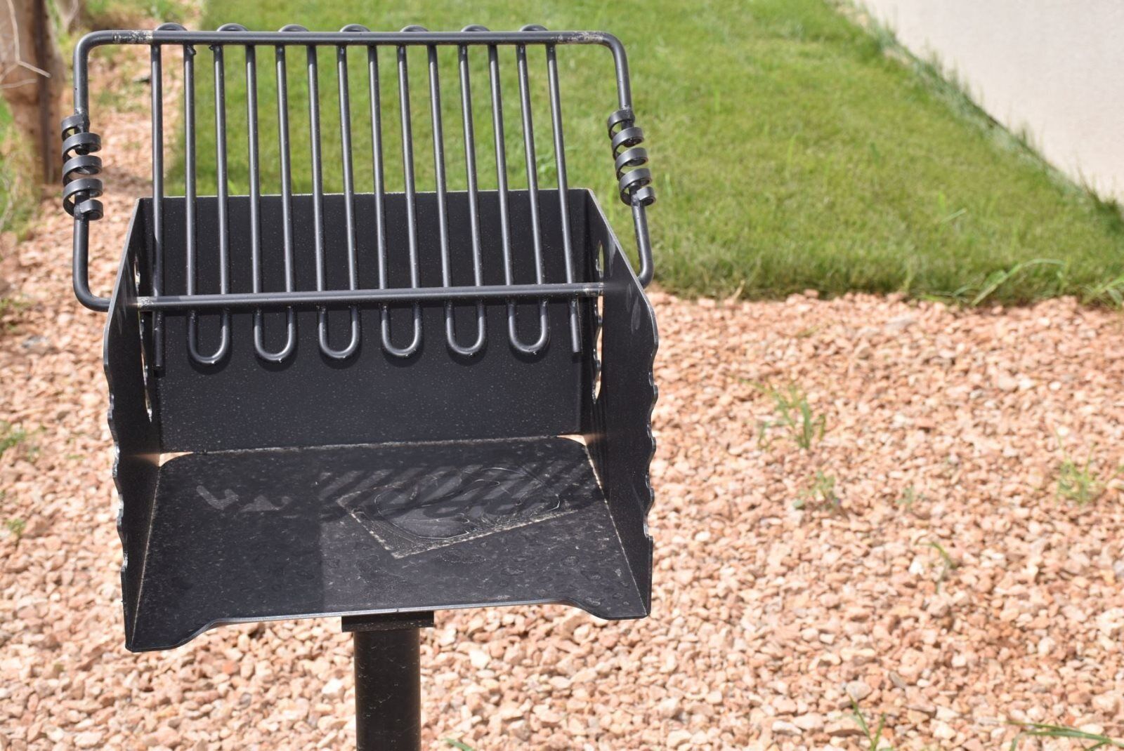 A black barbecue grill with a metal grate and a handle on the side, placed on a gravel surface with grass in the background.
