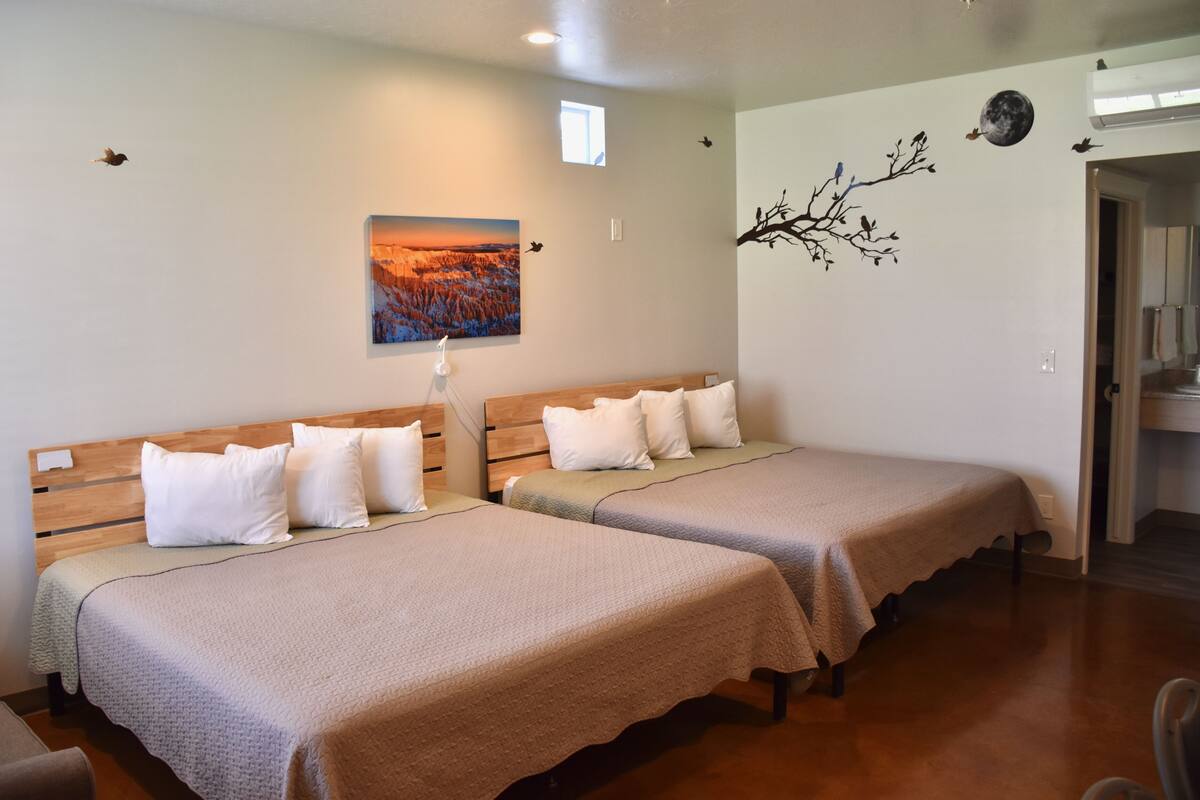 A hotel room with two single beds, a nightstand, a lamp, a painting of a sunset, and a wall decoration of a tree branch with birds.