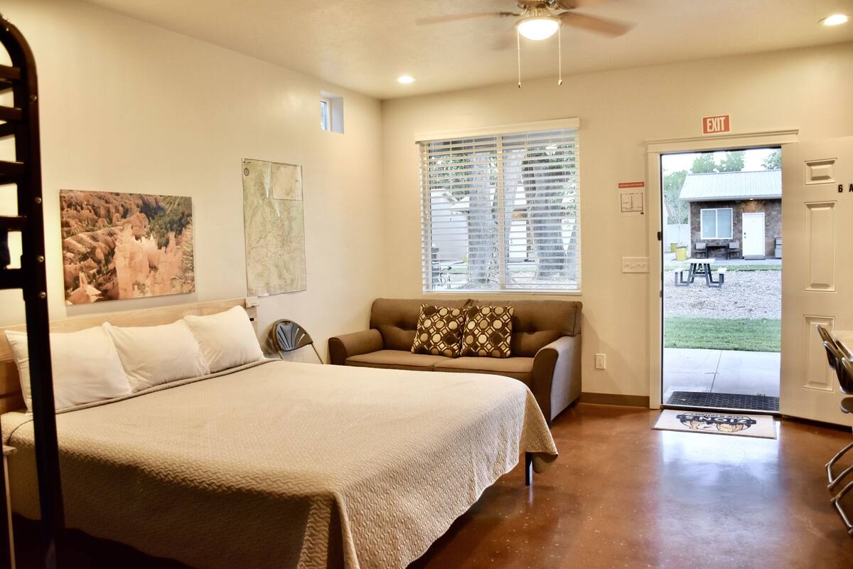 A hotel room with a bed, a sofa, a window with blinds, an exit sign, and a door leading outside.