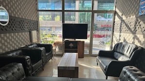 A modern living room with leather sofas, a flat-screen TV, and a large window offering a view of the street. - Asus Palmiye Otel (Adana)