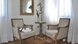 A room with two upholstered chairs, a round table with a lamp, a mirror, and a vase with flowers.