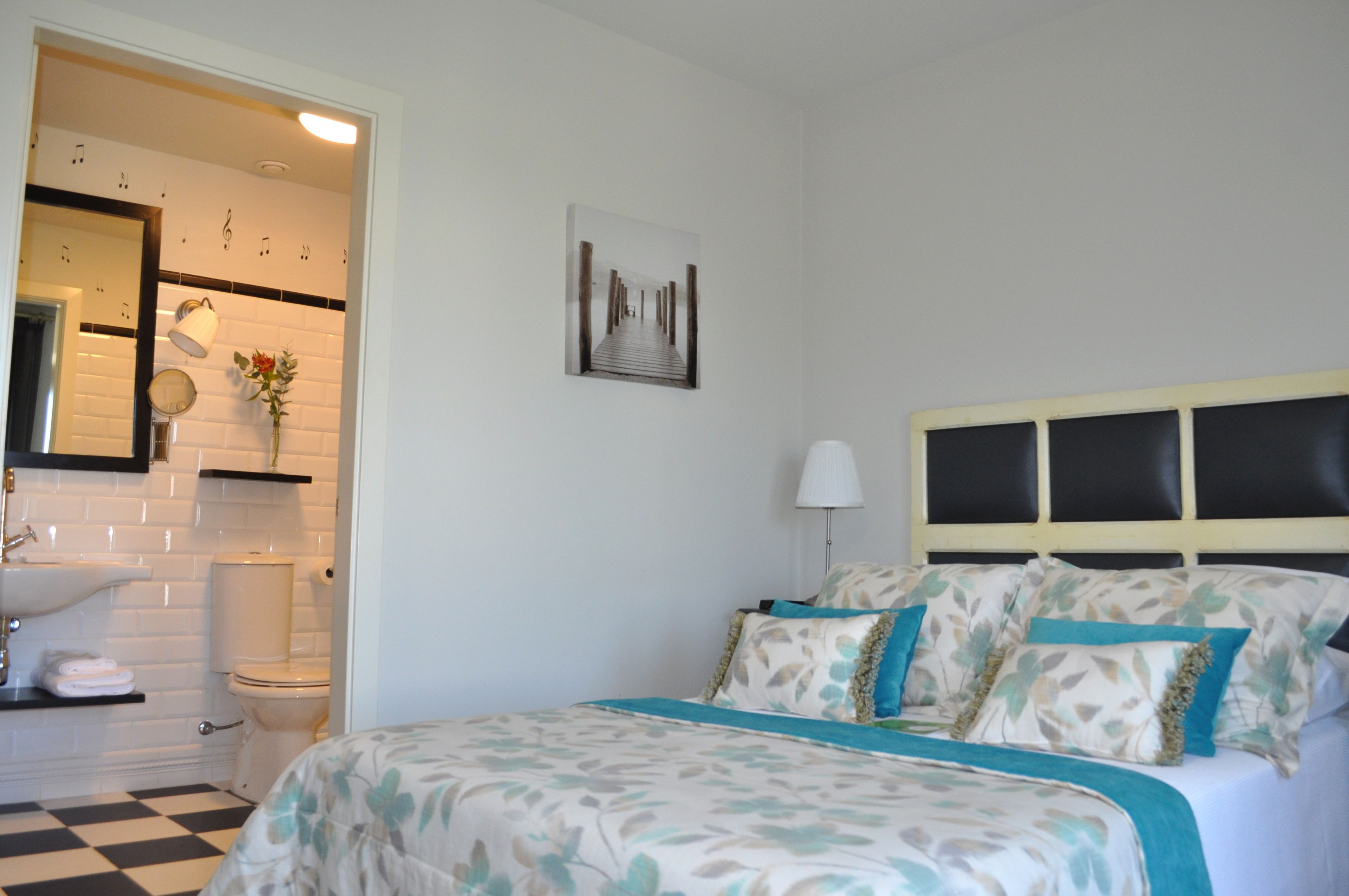 Ein Schlafzimmer mit Bett, Nachttisch, Lampe, einem Wandbild und einem Badezimmer mit Waschbecken und Toilette.