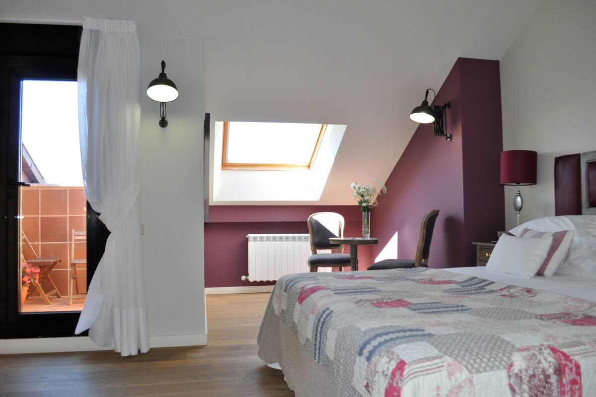 A bedroom with a large bed, a skylight, a chair, a table with a vase, and a view of an outdoor area.