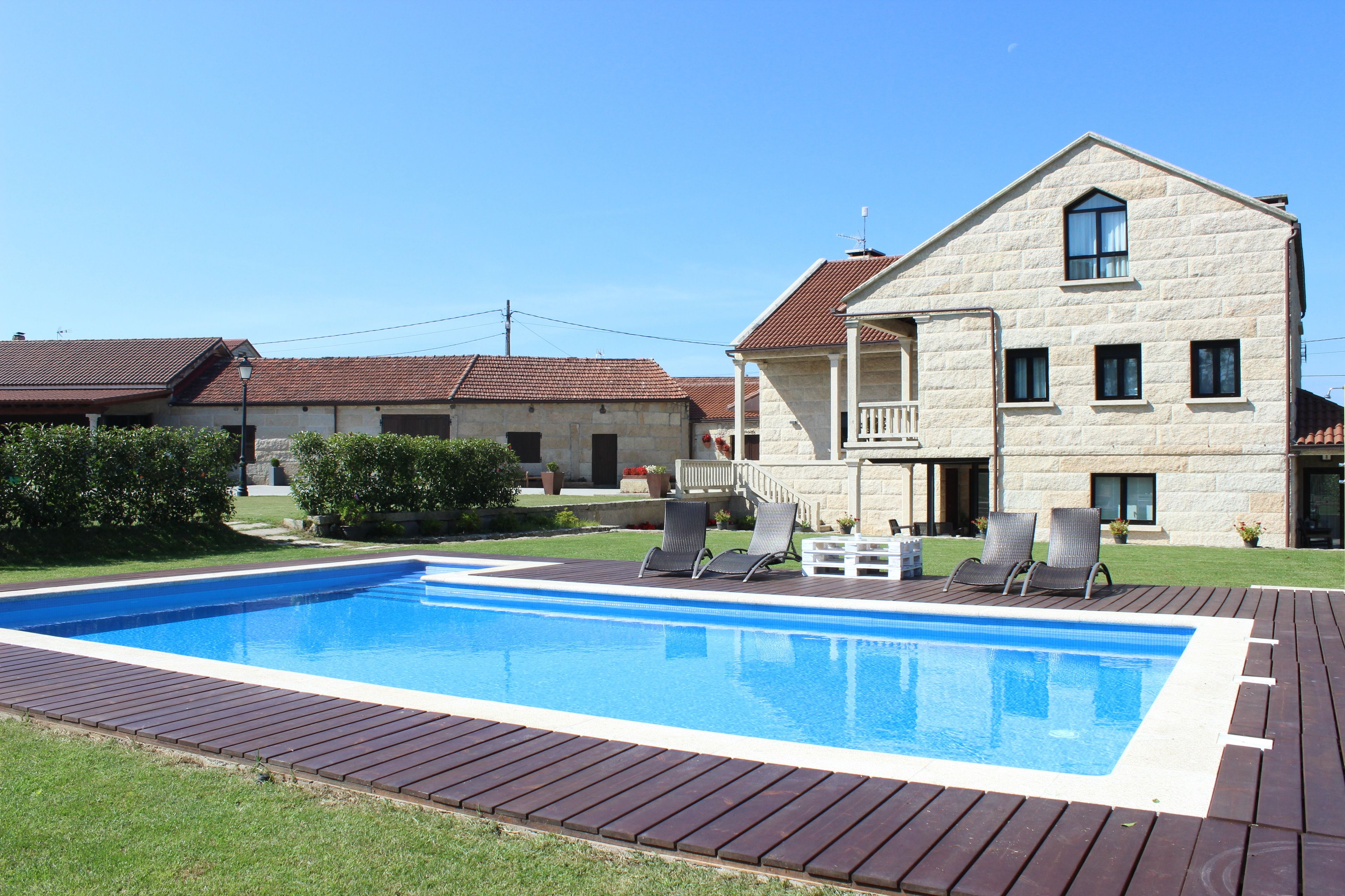 Una piscina con muelle de madera, rodeada por un césped cuidado y un edificio de piedra con balcón.