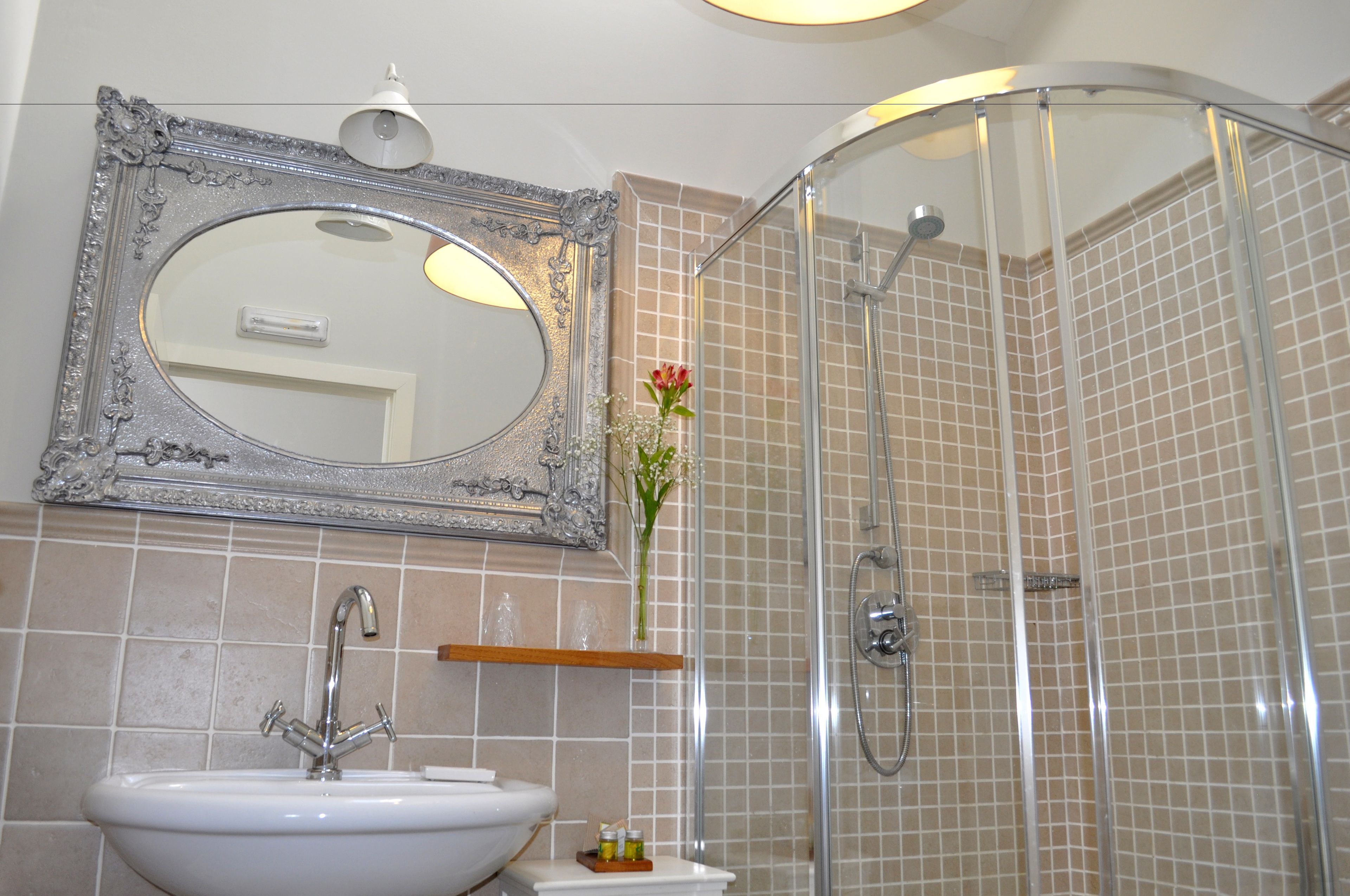A bathroom with a white sink, a mirror with an ornate frame, a shower with a glass door, and a vase with flowers on the counter.