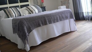 A neatly made bed with striped pillows, a bedside lamp, a vase with flowers, and a chair with a towel.