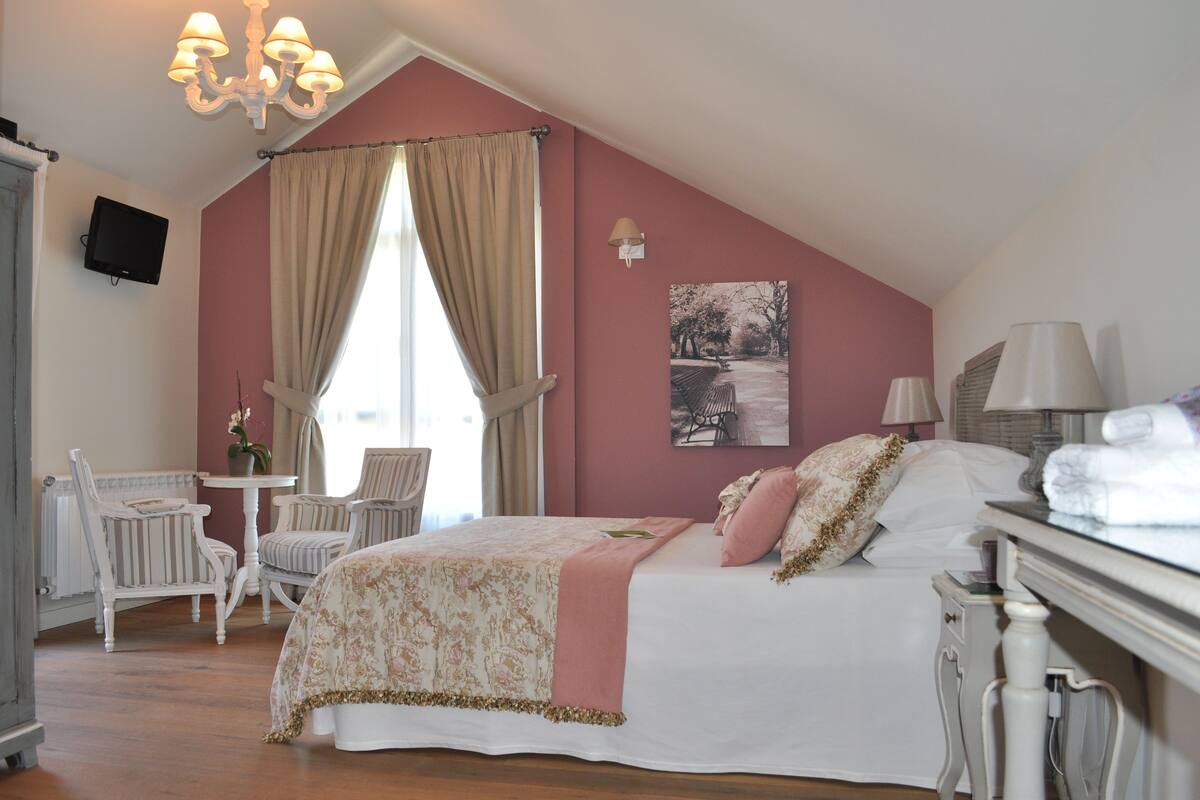 A bedroom with a sloped ceiling, a bed with floral bedding, a chair, a table, and a television mounted on the wall.