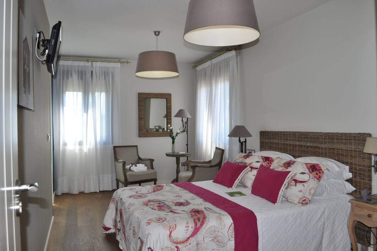 A bedroom with a bed, two chairs, a table, a mirror, and a TV.