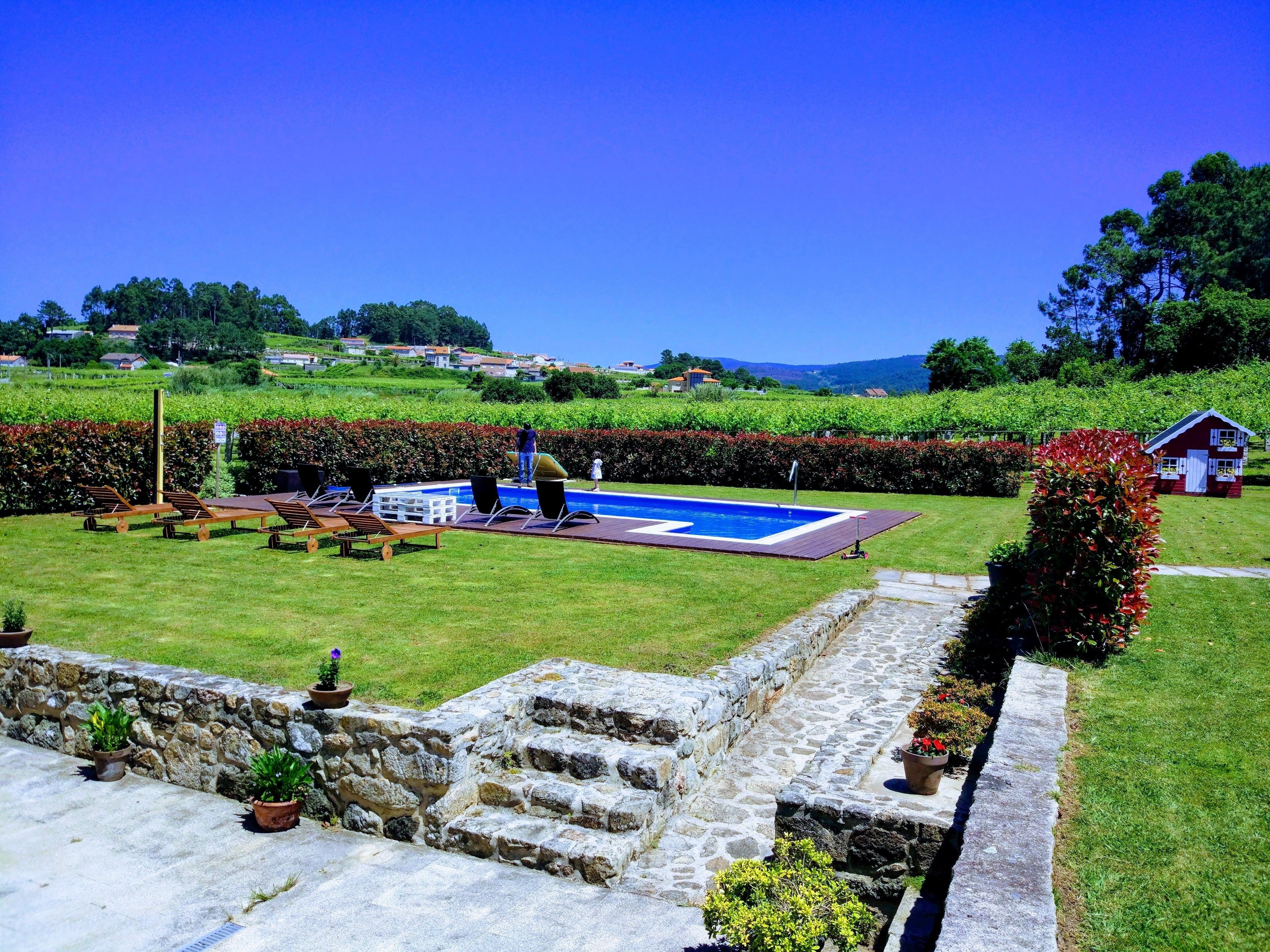 Un área de piscina con sillones de descanso, un muro de piedra y una casa pequeña al fondo.