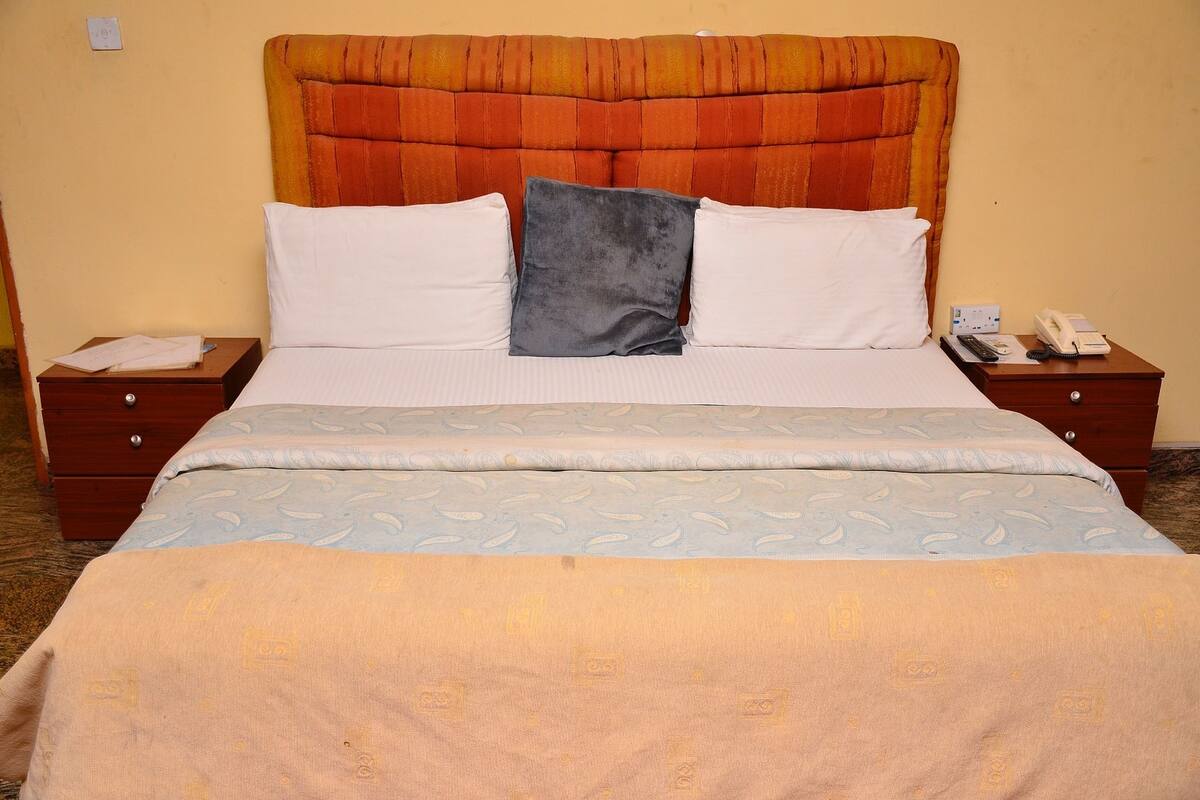 Una cama bien tendida con un edredón estampado, dos almohadas y un cabecero. Hay una mesita de noche con un teléfono y un control remoto.