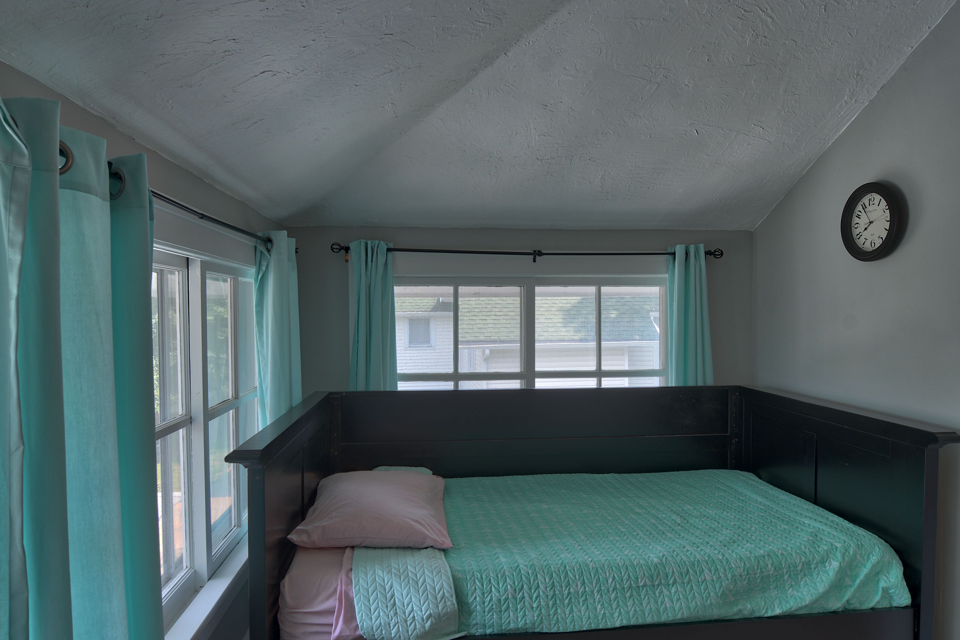 A small, single-bed room with a window, curtains, and a wall clock.