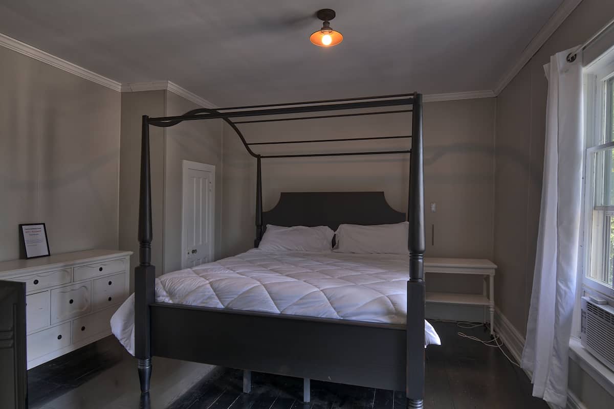 A four-poster bed with a dark frame, a white headboard, and a quilted white bedspread. There is a white bedside table with a lamp, a white dresser with drawers, and a window with white curtains.