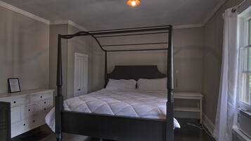 A four-poster bed with a dark frame, a white headboard, and a quilted white bedspread. There is a white bedside table with a lamp, a white dresser with drawers, and a window with white curtains.