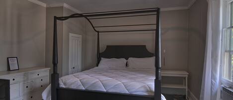 Una cama con dosel de estructura oscura, cabecera blanca acolchada y cubrecama blanco. Se observa una mesita de noche blanca con lámpara, un aparador blanco con cajones y una ventana con cortinas blancas.