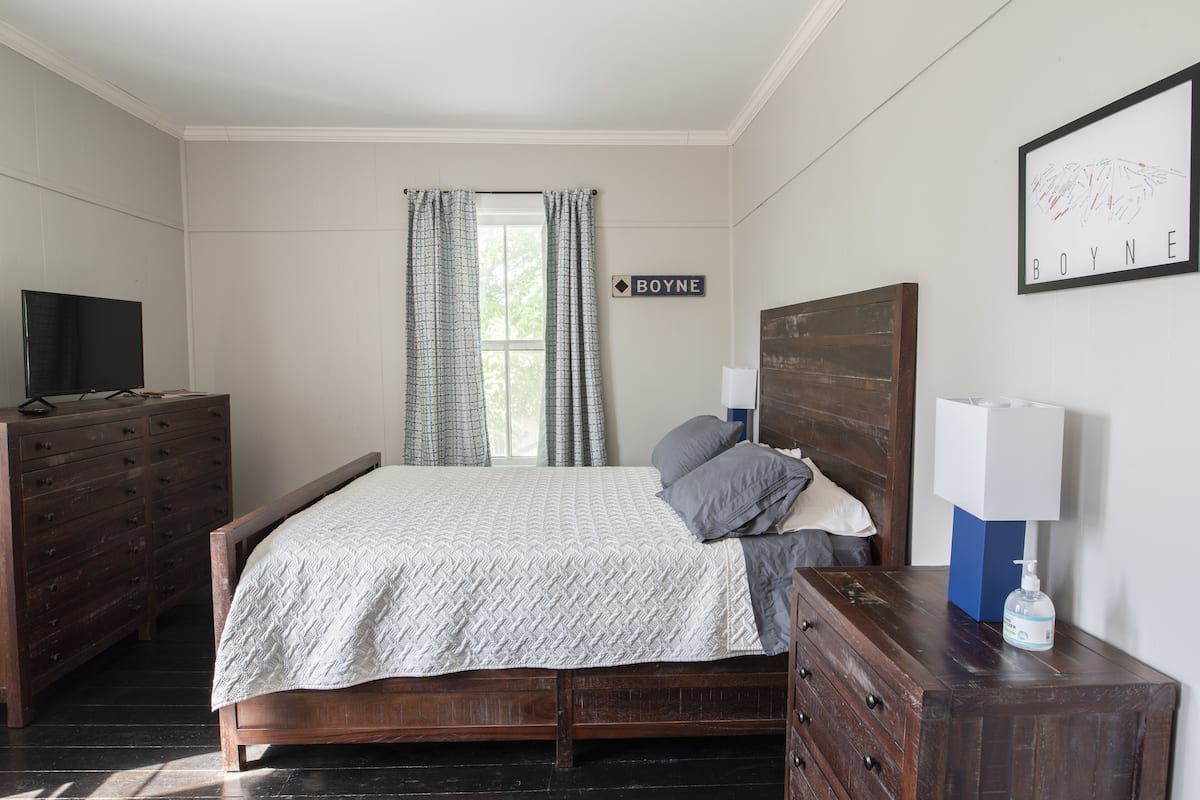 A bedroom with a bed, a dresser, a television, a window with curtains, and a wall sign.