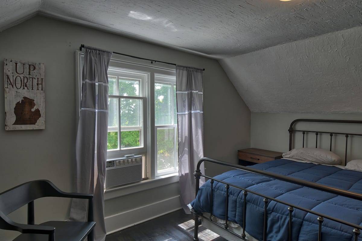 A bedroom with a metal bed frame, a window with curtains, a radiator, and a wall-mounted sign.