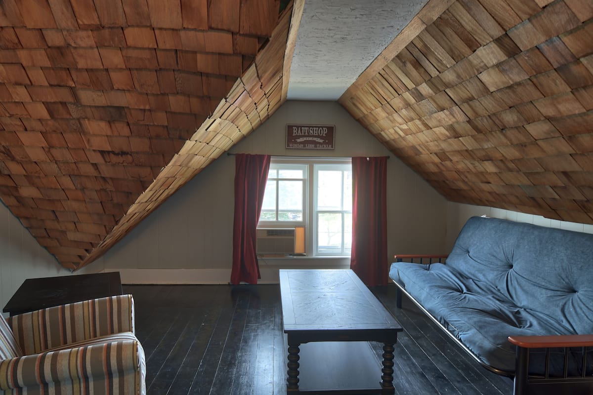 An attic room with a sloped wooden ceiling, a sofa, a striped chair, and a table. A sign above the window reads "BATT SHOP Worms Lies Tackle."