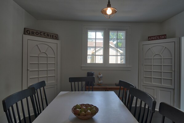 A dining area with a white table, four chairs, a basket of fruit, and two doors labeled "GROCERIES" and "APRÈS SKI".