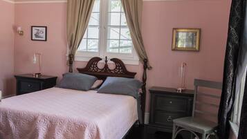 Un dormitorio con una cama, dos mesitas de noche, una silla, un ventanal con cortinas y dos cuadros en la pared.