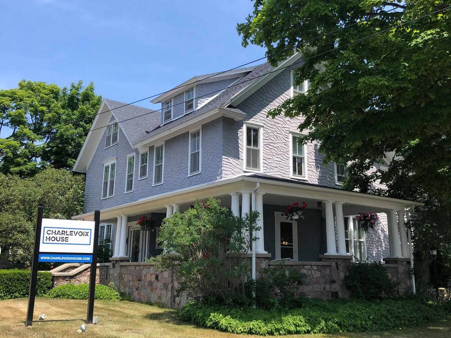 A two-story house with a stone foundation, a covered porch, and a sign indicating it is for sale.