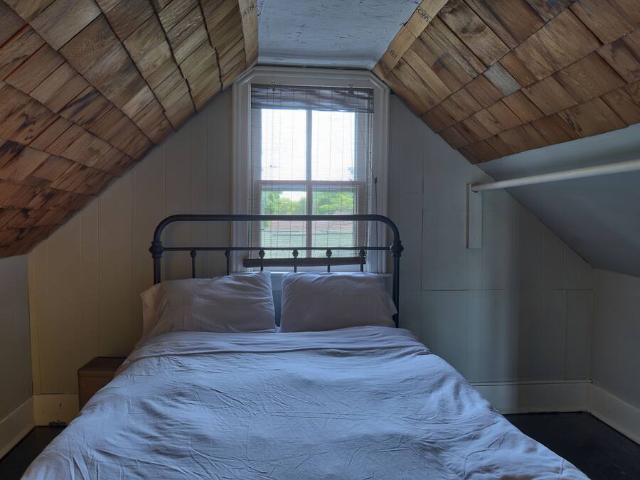 Un dormitorio con techo inclinado, una cama individual, un ventanal con persianas y un cabecero de madera.