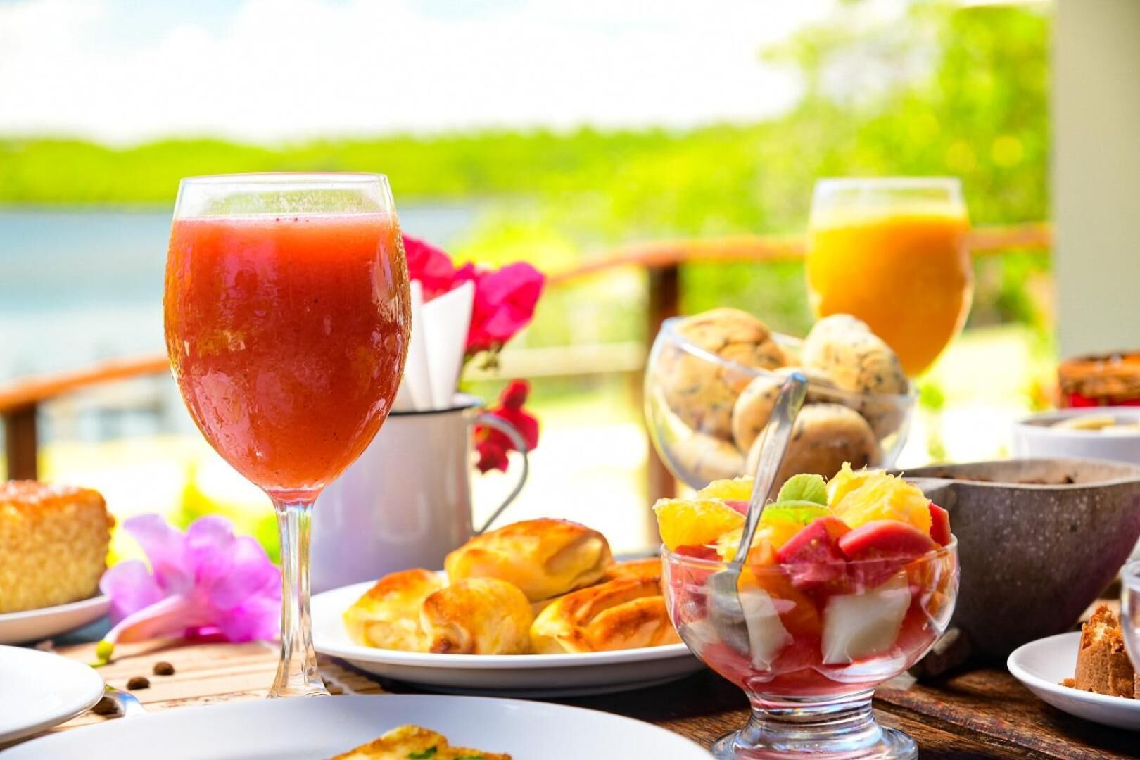 A breakfast setting with a glass of red juice, a bowl of fruit, pastries, and a view of a lake.