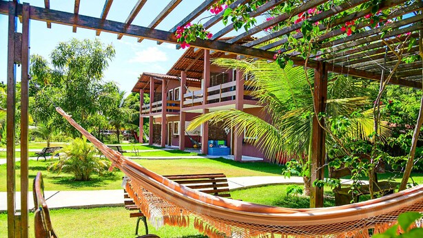 Una hamaca bajo una pérgola de madera con vistas a un edificio con varios balcones y un jardín cuidado.