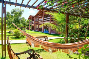 A hammock under a wooden pergola with a view of a building with multiple balconies and a well-maintained garden. - Pousada Porto Manguaba (Japaratinga)