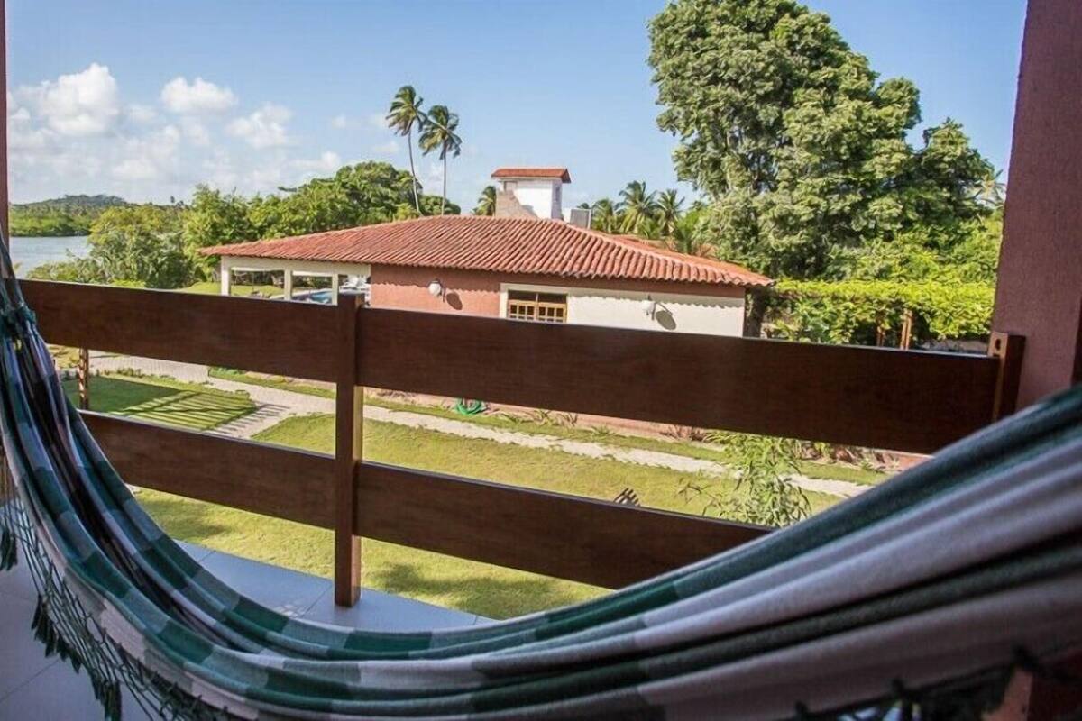 Uma rede numa varanda com vista para uma casa de telhado vermelho, rodeada por vegetação e um céu limpo.