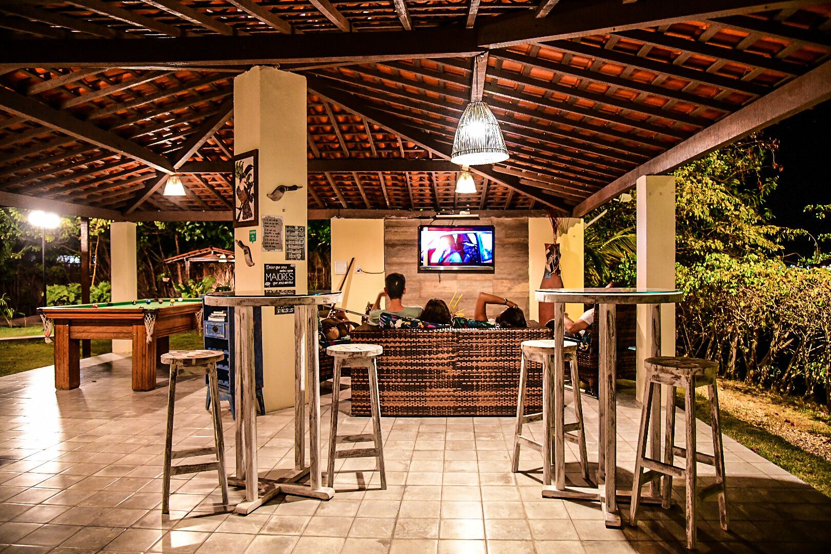 An outdoor bar area with a pool table, wooden stools, and a television screen.