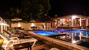 Un área de piscina nocturna con sillones de descanso, un bar y un restaurante.