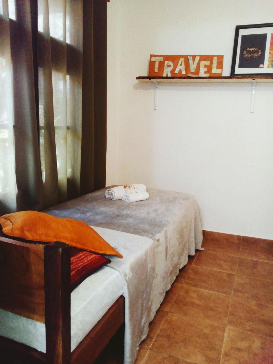 A small, single-bed room with a wooden chair, a towel on the bed, and a 'TRAVEL' sign on the wall.