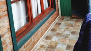 A room with a brick wall, a wooden-framed window with white curtains, a green door, and a tiled floor. - El Rinconcito (Puerto Iguazú)
