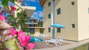 A modern building with a swimming pool, outdoor seating, and a covered area with a blue umbrella. - Flats Vieiras do Maragogi (Maragogi)