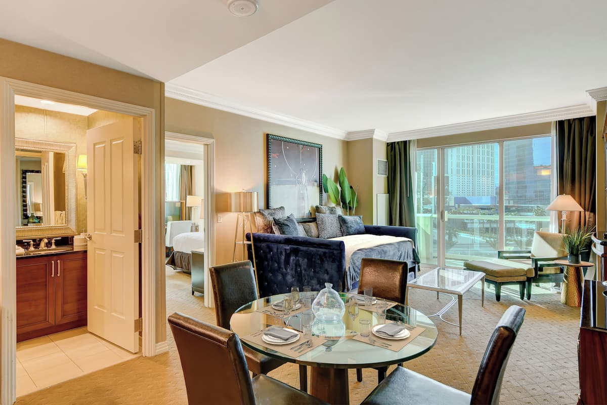 A hotel room with a dining area, a bed with a blue headboard, a large window, and a view of the city.