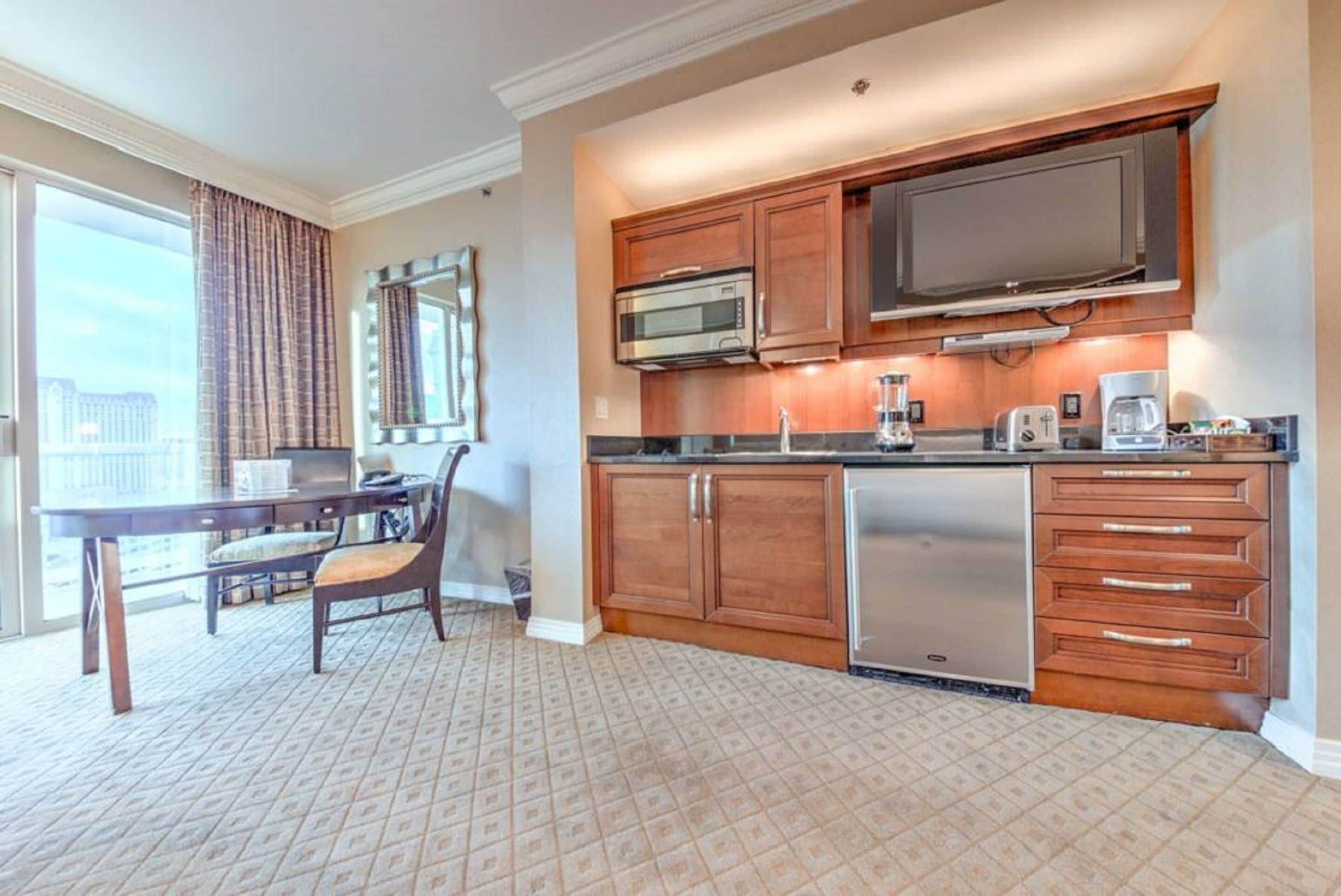 A hotel room with a kitchenette, a dining area with a table and chairs, and a large window with curtains.