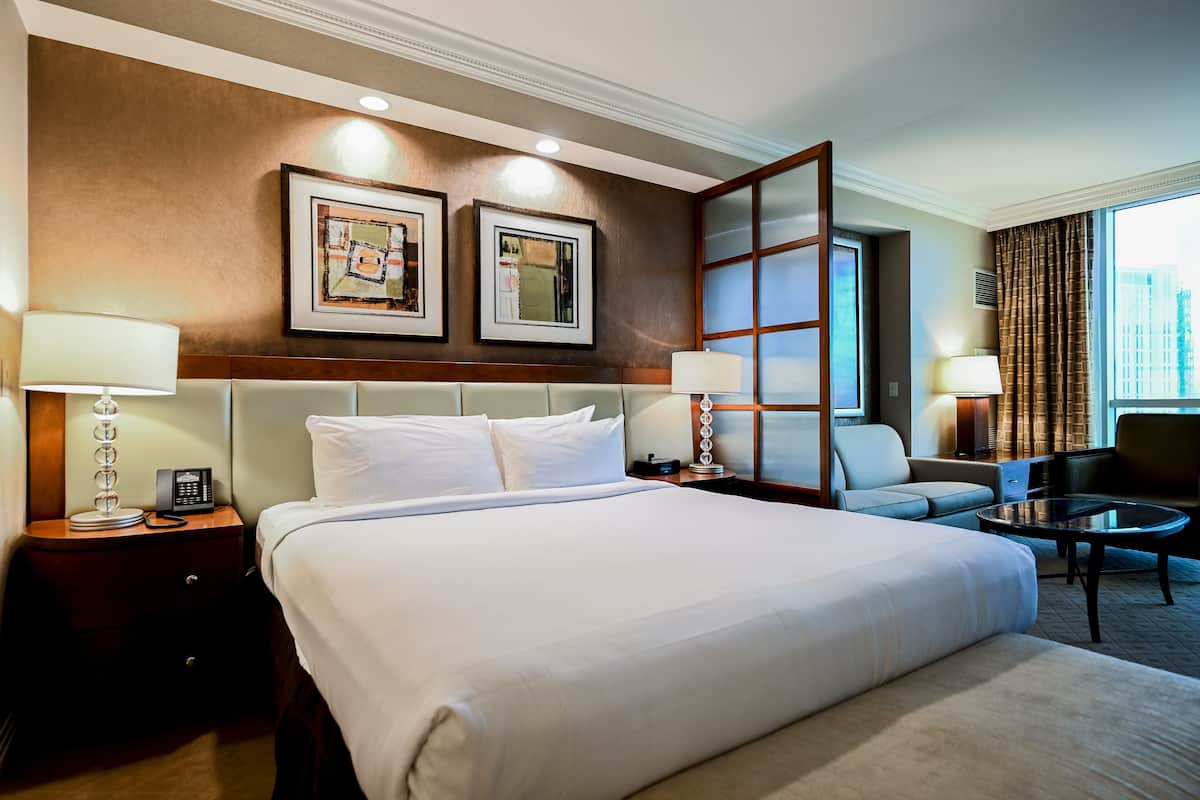 A hotel room with a large bed, two bedside tables with lamps, a sofa, a chair, and a large window with curtains.