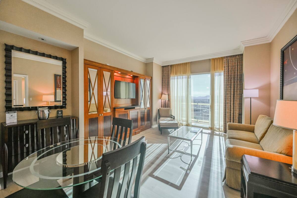 A hotel room with a dining area, a sofa, a TV, and a large window with curtains.