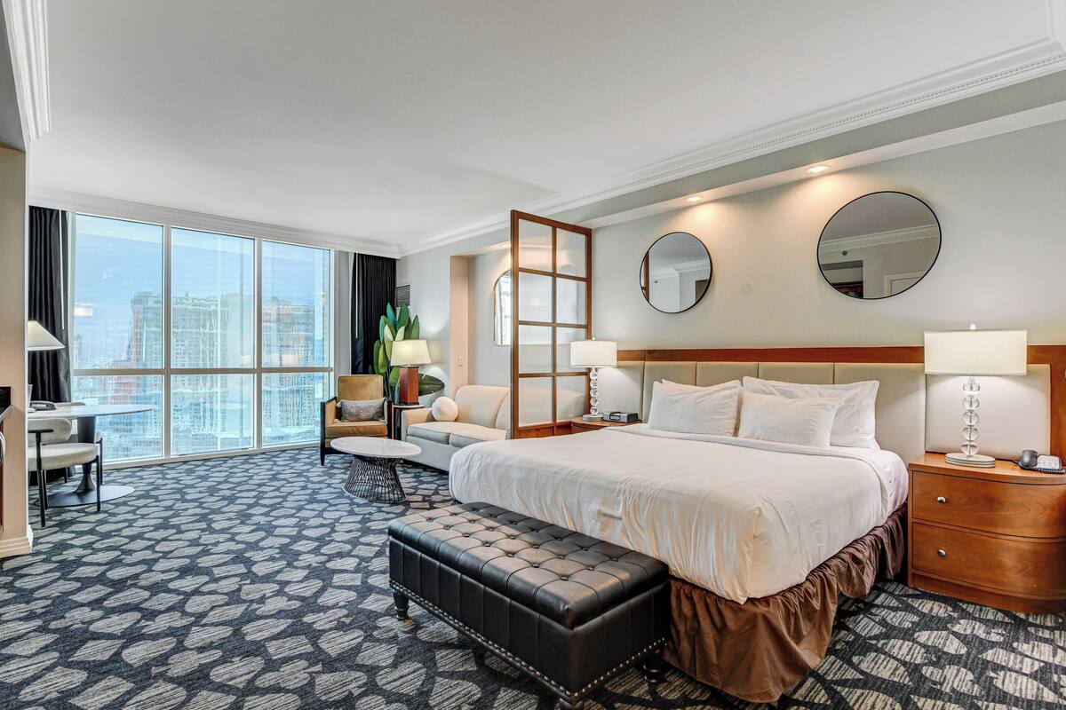 A hotel room with a large bed, a sitting area with a sofa and armchair, a small table, and a large window offering a city view.