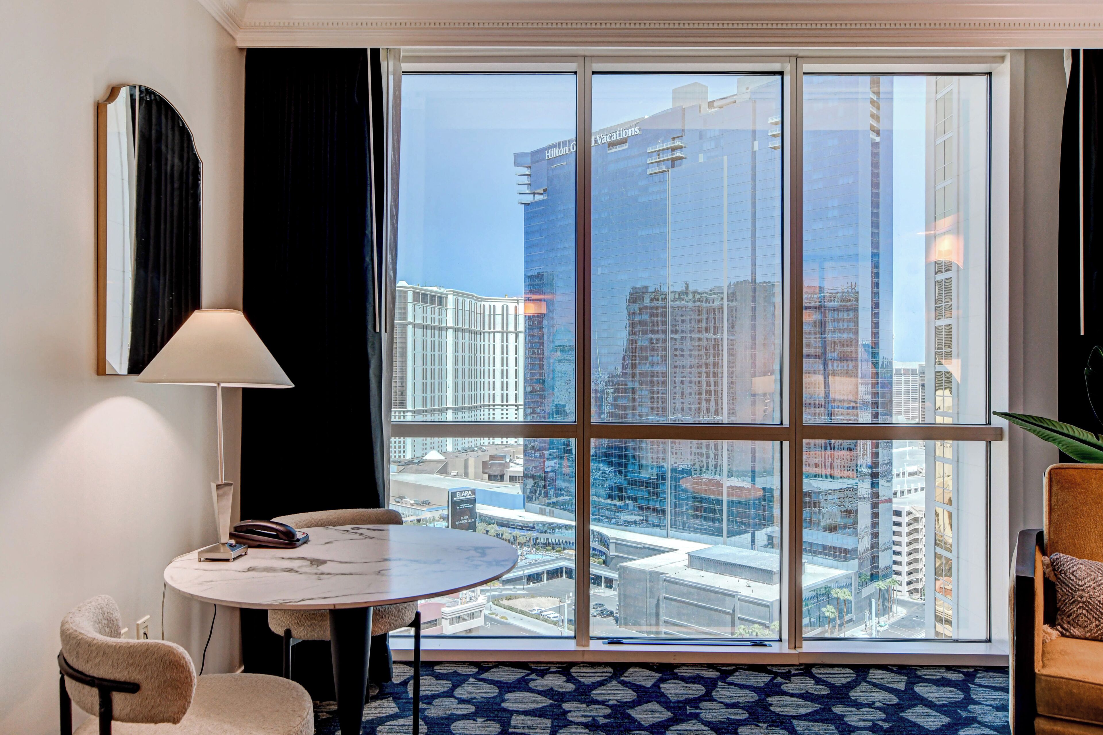 A hotel room with a large window offering a city view, a round table, a chair, a lamp, and a plant.