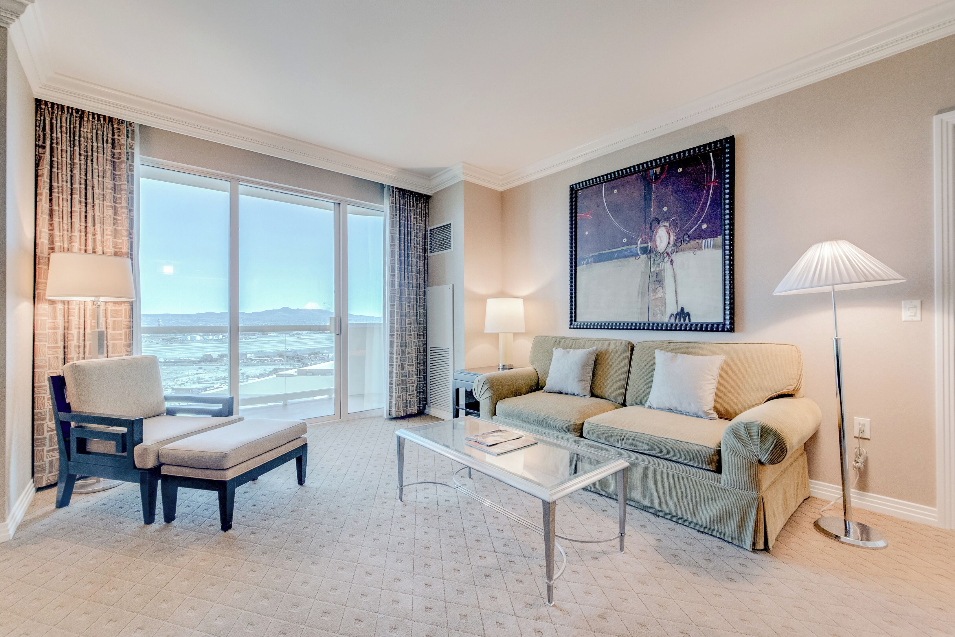 Une chambre d’hôtel moderne dotée d’une grande fenêtre, d’un canapé, d’un fauteuil, d’une table basse et d’un lampadaire.